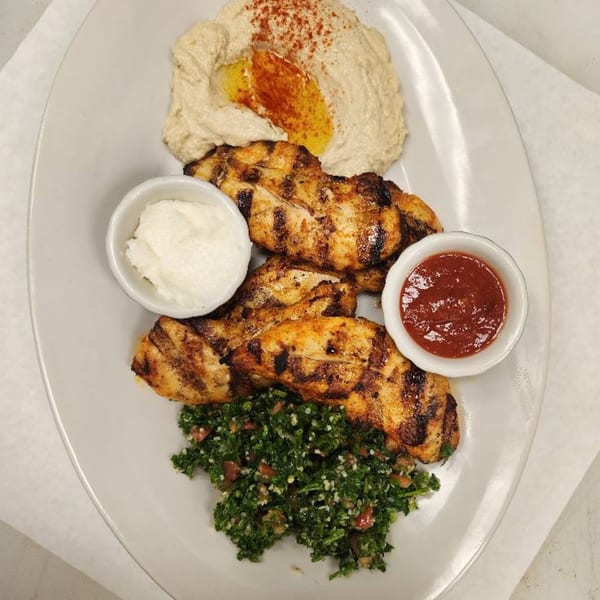 Grilled chicken with hummus, tabbouleh, garlic sauce, and red sauce on a white plate.