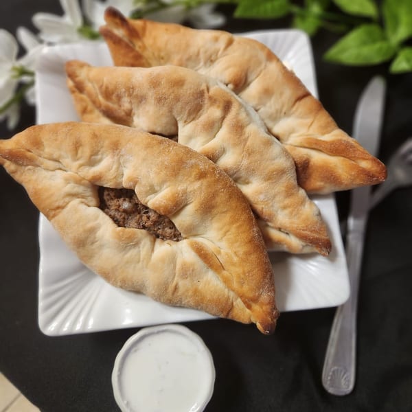 Four golden-brown meat pies, one open to show filling, on a white plate with a side of sauce.